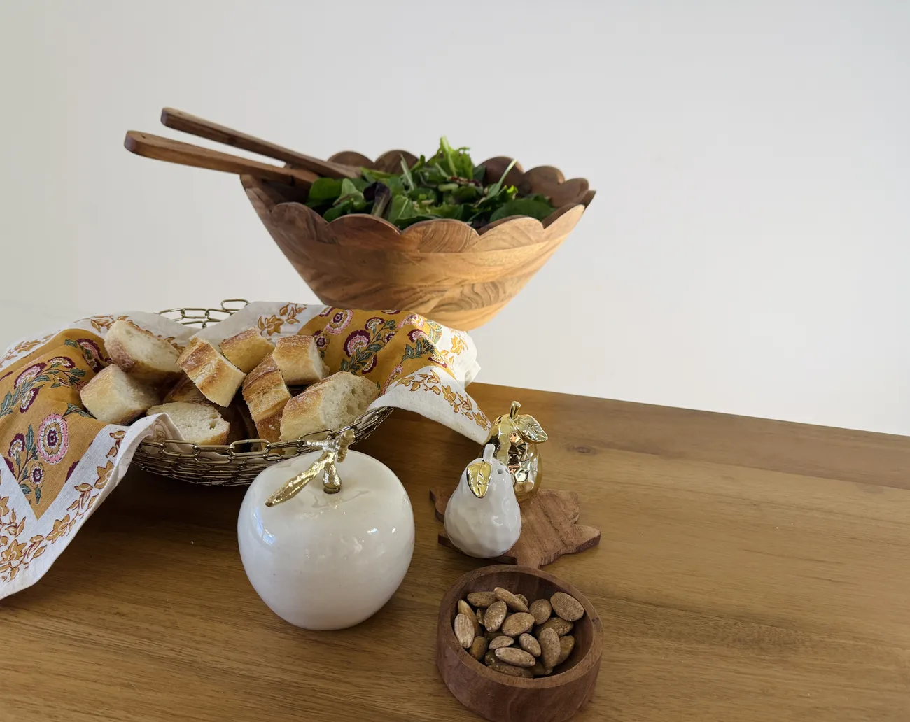 A scalloped wooden salad bowl of dressed leaves beside a wire basket of sliced baguette and cream ceramic fruit